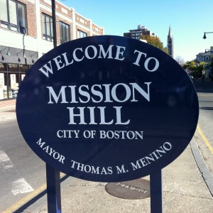 The “Welcome to Mission Hill” sign in Brigham Circle at Tremont Street and Huntington Avenue still bears the name of Mayor Thomas Menino. (Gazette Photo by Rebeca Oliveira)