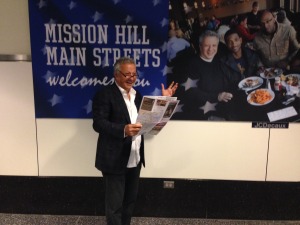 Michel Soltani, vice president of the Mission Hill Main Streets board and owner of the Mission Bar and Grill, poses in front of an advertisement for Mission Hill in Logan Airport’s Terminal A last month. The advertising firm JCDecaux donated three large ads at the airport to MHMS as part of a promotion seeking to draw tourists to Boston’s local businesses. (Photo by Richard Rouse/MHMS)